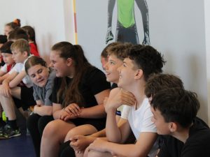 Uczniowie siedzący na ławce w hali sportowej obserwują przebieg turnieju piłki ręcznej rozgrywanego z okazji Dnia Dziecka.