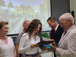 Uczennica szkoły podstawowej w Ostaszewie odbiera pamiątkowy dyplom z rąk przedstawiciela samorządu podczas Gali Absolwentów Gminy Łysomice. Uczennicy towarzyszą rodzice również przyjmujący gratulacje.