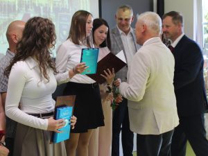 Uczennica szkoły podstawowej w Ostaszewie odbiera pamiątkowy dyplom z rąk przedstawiciela samorządu podczas Gali Absolwentów Gminy Łysomice. Uczennicy towarzyszą rodzice również przyjmujący gratulacje.