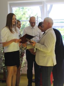 Uczennica szkoły podstawowej w Ostaszewie odbiera pamiątkowy dyplom z rąk przedstawiciela samorządu podczas Gali Absolwentów Gminy Łysomice. Uczennicy towarzyszą rodzice również przyjmujący gratulacje.
