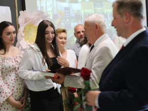 Uczennica szkoły podstawowej w Ostaszewie odbiera pamiątkowy dyplom z rąk przedstawiciela samorządu podczas Gali Absolwentów Gminy Łysomice. Uczennicy towarzyszą rodzice również przyjmujący gratulacje.