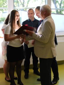 Uczennica szkoły podstawowej w Ostaszewie odbiera pamiątkowy dyplom z rąk przedstawiciela samorządu podczas Gali Absolwentów Gminy Łysomice. Uczennicy towarzyszą rodzice również przyjmujący gratulacje.