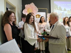 Uczennica szkoły podstawowej w Ostaszewie odbiera pamiątkowy dyplom z rąk przedstawiciela samorządu podczas Gali Absolwentów Gminy Łysomice. Uczennicy towarzyszą rodzice również przyjmujący gratulacje.