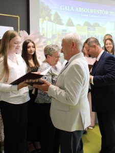 Uczennica szkoły podstawowej w Ostaszewie odbiera pamiątkowy dyplom z rąk przedstawiciela samorządu podczas Gali Absolwentów Gminy Łysomice. Uczennicy towarzyszą rodzice również przyjmujący gratulacje.