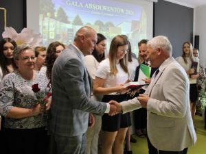 Uczennica szkoły podstawowej w Ostaszewie odbiera pamiątkowy dyplom z rąk przedstawiciela samorządu podczas Gali Absolwentów Gminy Łysomice. Uczennicy towarzyszą rodzice również przyjmujący gratulacje.