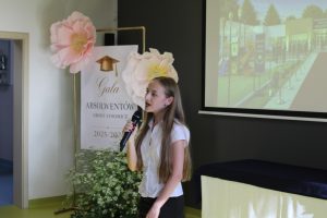 Uczennica wykonuje utwór wokalny podczas części artystycznej Gali Absolwentów. W tle znajduje się dekoracja wydarzenia oraz ekran z prezentacją.