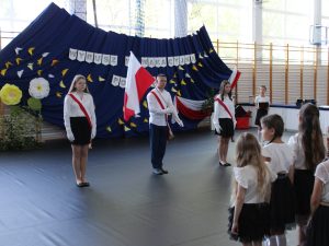 Poczet flagowy szkoły stoi na środku sali gimnastycznej podczas uroczystego zakończenia roku szkolnego. W tle widoczna dekoracja z napisem „Wyrusz w wakacyjną podróż”.