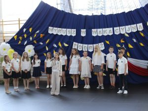 Uczniowie klasy III prezentują program artystyczny. Na pierwszym planie dziewczynka śpiewa do mikrofonu, a pozostali uczniowie stoją za nią w półkolu.