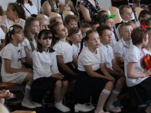 Dzieci zgromadzone na widowni z zainteresowaniem oglądają część artystyczną uroczystości. Wśród uczniów widoczne są kwiaty przygotowane dla nauczycieli.