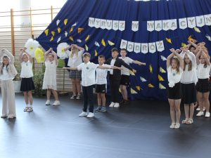 Uczniowie klasy III prezentują choreografię przygotowaną na zakończenie roku szkolnego. Dzieci stoją w różnych układach na tle dekoracji.