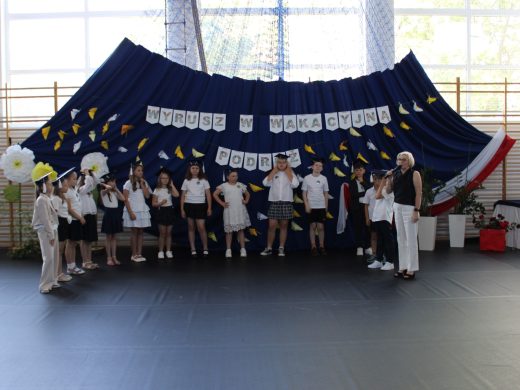 Dyrektor szkoły przemawia do uczniów klasy III podczas uroczystości zakończenia pierwszego etapu edukacyjnego. Uczniowie występują w symbolicznych biretach.