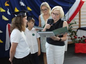 Uczennica klasy III odbiera świadectwo i książkę z rąk dyrektor szkoły podczas uroczystego zakończenia roku szkolnego.