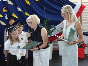 Dyrektor szkoły wręcza kolejnemu uczniowi świadectwo ukończenia klasy III i pamiątkową książkę. Obok stoi uśmiechnięta wychowawczyni dziewczynki.