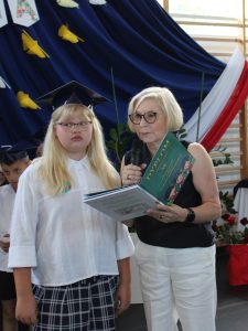 Uczennica klasy III odbiera świadectwo oraz książkę w obecności wychowawczyni i pozostałych uczniów.