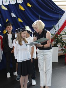 Uczennica w birecie odbiera świadectwo oraz książkę na zakończenie pierwszego etapu edukacyjnego.