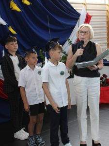 Dyrektor szkoły wręcza świadectwo uczniowi klasy III, który stoi w birecie obok swoich kolegów i koleżanek.