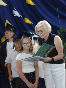 Uczennica klasy trzeciej odbiera świadectwo i pamiątkową książkę z rąk dyrektor szkoły podczas uroczystego zakończenia roku szkolnego.