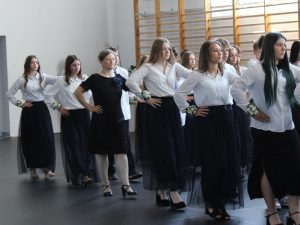 Uczniowie klasy ósmej oraz wychowawczyni wykonują tradycyjny taniec podczas uroczystości zakończenia roku szkolnego na sali gimnastycznej. Uczestnicy ubrani są odświętnie, a dziewczęta mają przypięte białe kwiaty na nadgarstkach.