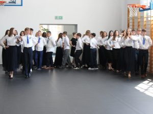 Pary uczniów klas ósmych ustawione w w półokręgu podczas wykonywania poloneza na sali gimnastycznej.