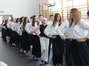 Uczniowie klasy ósmej wraz z wychowawczynią i panią dyrektor tańczą poloneza ustawieni w pary podczas uroczystości zakończenia roku szkolnego. Wszyscy stoją w jednym rzędzie na sali gimnastycznej.