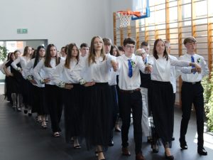 Pary uczniów klas ósmych tańczą poloneza podczas uroczystości zakończenia roku szkolnego. Uczniowie ustawieni są w cztery kolumny i wykonują zsynchronizowany układ.