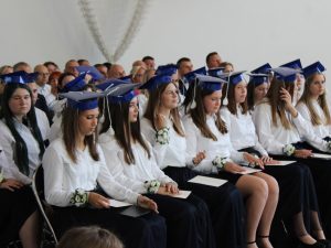 Absolwenci szkoły siedzą w pierwszych rzędach podczas uroczystości zakończenia roku szkolnego. Na głowach mają granatowe birety, a w dłoniach trzymają dokumenty i pamiątki.