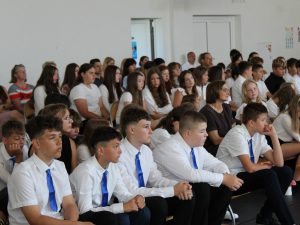 Uczniowie oraz rodzice zgromadzeni na sali gimnastycznej słuchają przemówień i podsumowania roku szkolnego podczas uroczystości zakończenia roku.
