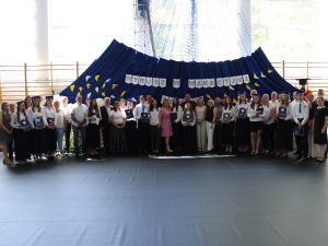 Pamiątkowe zdjęcie absolwentów, rodziców, nauczycieli i zaproszonych gości wykonane po zakończeniu uroczystości. Uczestnicy pozują na tle dekoracji z hasłem wakacyjnym.
