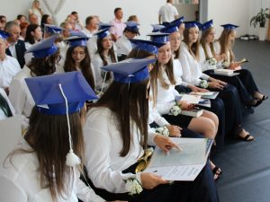 Absolwenci w granatowych biretach oglądają otrzymane świadectwa i nagrody podczas zakończenia roku szkolnego. W tle widoczni są rodzice i pozostali goście uroczystości.