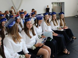 Uczennice klasy ósmej siedzą podczas uroczystości zakończenia roku szkolnego. Ubrane są w białe koszule i granatowe stroje galowe, mają na głowach niebieskie birety. W dłoniach trzymają świadectwa, dyplomy i upominki. W tle widoczni są rodzice oraz zaproszeni goście obserwujący przebieg uroczystości.