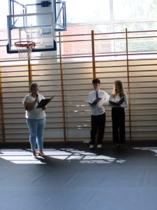 Przedstawicielka rodziców absolwentów szkoły przemawia do uczestników uroczystości, trzymając mikrofon i tekst wystąpienia. Obok stoją uczniowie prowadzący akademię, ubrani w stroje galowe i trzymający przygotowane scenariusze.