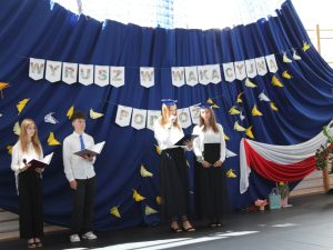 Uczniowie prowadzący część artystyczną wraz z dwoma przedstawicielkami klasy ósmej występują na tle dekoracji z napisem „Wyrusz w wakacyjną podróż”. Na granatowej kurtynie zawieszono papierowe żurawie i biało-czerwoną dekorację nawiązującą do barw narodowych.