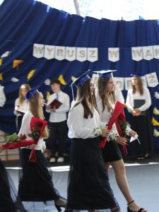 Absolwentki w niebieskich biretach przechodzą przez salę gimnastyczną, niosąc czerwone róże. W tle widoczni są uczniowie prowadzący uroczystość oraz wakacyjna dekoracja przygotowana na zakończenie roku szkolnego.