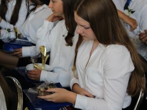 Zbliżenie na uczennicę siedzącą wśród absolwentów. Trzyma w dłoniach pamiątkowy upominek i trofeum wręczone podczas uroczystości zakończenia szkoły podstawowej.