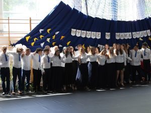 Uczniowie klasy ósmej stoją razem na tle dekoracji z napisem „Wyrusz w wakacyjną podróż”. Absolwenci obejmują się ramionami, tworząc wspólny krąg podczas części artystycznej przygotowanej na zakończenie nauki w szkole podstawowej.