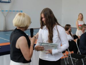 Dyrektor szkoły wręcza uczennicy klasy szóstej świadectwo oraz nagrodę książkową. W tle siedzą nauczyciele i rodzice uczestniczący w uroczystości zakończenia roku szkolnego.
