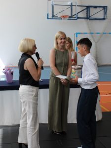 Dyrektor szkoły oraz wychowawczyni wręczają uczniowi klasy szóstej świadectwo i nagrody za osiągnięcia edukacyjne. Uczeń odbiera wyróżnienie podczas oficjalnej części uroczystości.