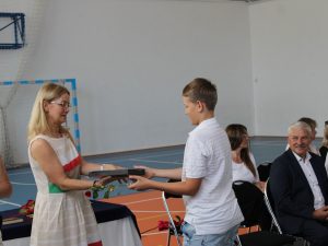 Wychowawczyni wręcza uczniowi nagrodę książkową oraz pamiątkowy upominek. W pierwszym rzędzie siedzą nauczyciele, wśród nich przedstawiciele władz samorządowych uczestniczący w uroczystości.