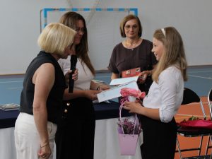 Dyrektor szkoły i nauczycielki wręczają uczennicy klasy czwartej świadectwo z wyróżnieniem oraz nagrody książkowe. Uczennica trzyma także koszyczek z kwiatami przygotowany dla nauczycieli.