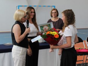 Uczennica klasy czwartej odbiera świadectwo i nagrody podczas uroczystości zakończenia roku szkolnego. W dłoniach trzyma kolorowy bukiet kwiatów wręczony nauczycielom w podziękowaniu za lata nauki.