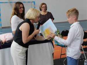 Dyrektor szkoły wręcza uczniowi klasy czwartej świadectwo oraz nagrody książkowe. W tle znajdują się wychowawczynie chłopca.