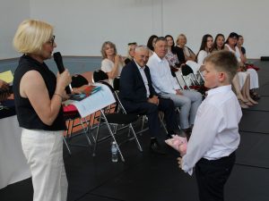 Dyrektor szkoły zwraca się do ucznia klasy czwartej odbierającego nagrody, podczas gdy nauczyciele oraz zaproszeni goście obserwują przebieg ceremonii.