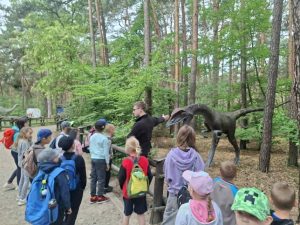 Dzieci z oddziału przedszkolnego podczas zwiedzania ścieżki edukacyjnej w JuraParku w Solcu Kujawskim. Przewodnik opowiada o modelu prehistorycznego dinozaura ustawionego wśród drzew.