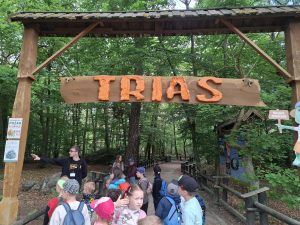 Grupa dzieci wraz z przewodnikiem stoi pod drewnianą bramą z napisem „TRIAS”, rozpoczynając zwiedzanie ścieżki edukacyjnej poświęconej dinozaurom.