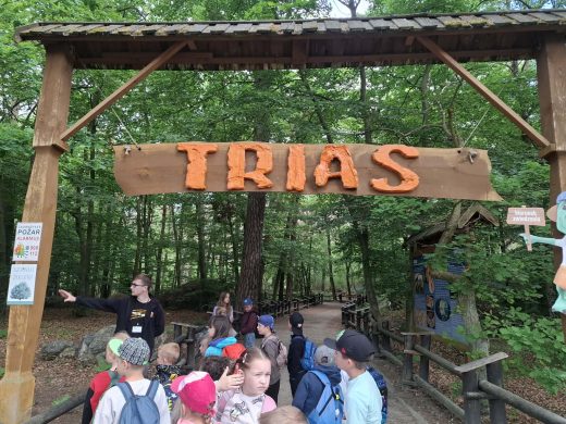 Grupa dzieci wraz z przewodnikiem stoi pod drewnianą bramą z napisem „TRIAS”, rozpoczynając zwiedzanie ścieżki edukacyjnej poświęconej dinozaurom.