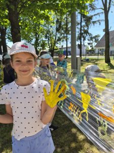 Uczennica prezentuje dłoń pomalowaną żółtą farbą podczas wspólnego tworzenia plastycznej instalacji na folii rozciągniętej między drzewami. W tle widoczni są inni uczniowie malujący kolorowe wzory na szkolnym terenie.