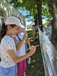 Uczennice malują kolorowe wzory na przezroczystej folii rozciągniętej między drzewami. Zajęcia plastyczne odbywają się na świeżym powietrzu w cieniu szkolnych drzew.