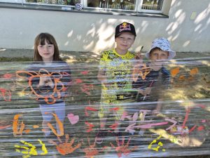 Troje uczniów stoi przy przezroczystej folii ozdobionej kolorowymi rysunkami i odciskami dłoni. Dzieci uczestniczą w plenerowych zajęciach plastycznych zorganizowanych podczas Dnia Sportu i Zdrowia.
