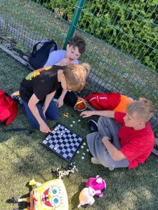 Uczniowie klas IV–VIII siedzą na boisku szkolnym wokół planszy do warcabów. Podczas pikniku z okazji Dnia Sportu i Zdrowia grają w gry planszowe i spędzają wspólnie czas na świeżym powietrzu.
