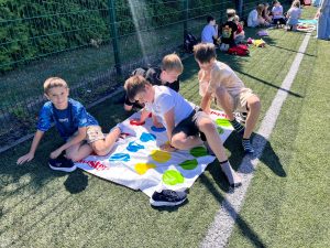 Grupa uczniów uczestniczy w grze integracyjnej na macie Twister rozłożonej na boisku szkolnym podczas obchodów Dnia Sportu i Zdrowia.